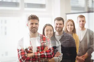 Five people stand in a row indoors, facing the camera and smiling. The group includes both men and women, and the setting appears to be an office with large windows.