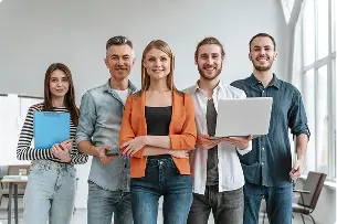 A diverse group of five people standing indoors, smiling confidently. Casual attire, one holds a clipboard, another a laptop. Bright, modern office setting.