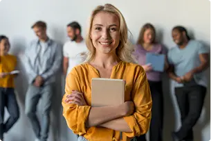 A woman in a yellow shirt stands holding a notebook and smiling at the camera, with a group of people talking in the background.