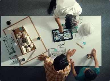 Three people collaborate at a table with architectural plans, a scale model, and digital floor plans. The scene conveys teamwork and creative focus.