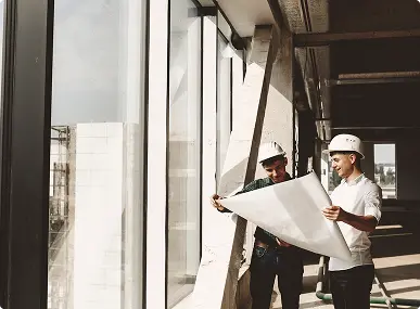 Two construction workers in hard hats examine blueprints by a large window inside a building under construction, conveying focus and collaboration.